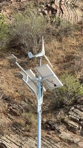 A stationary, eye-level shot shows a hybrid solar and wind-powered streetlight on a pole in a natural, hilly environment. The streetlight has two LED lamps facing outwards and upwards. Mounted above the lamps are two solar panels, angled to catch sunlight, and a small wind turbine with three blades positioned at the very top. The pole is anchored to the ground, but the base is not visible. Behind the streetlight, a steep, rocky hillside is covered with dry brush and sparse vegetation. The rock formations are layered and appear eroded, suggesting a dry, arid climate. The lighting indicates it is daytime. There are no people or animals visible in the frame, and no actions are taking place beyond the natural movement of the wind turbine blades, which appear to be slowly rotating.