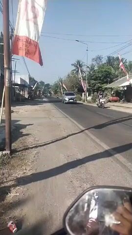 The video shows a road with flags and decorations on the sides, suggesting a festive or celebratory occasion. The flags are red and white, common colors in many national flags, including Indonesia's. A silver minivan is driving down the road, and a person is riding a motorcycle on the right side of the road. The scene appears to be in a rural or suburban area, with trees and houses visible in the background. The video is shot from the perspective of someone riding a motorcycle, as evidenced by the reflection in the rearview mirror.