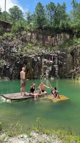 Four individuals are on a raft in a quarry lake. One man stands on the left of the raft, while three others sit. The man standing is wearing shorts. The two sitting on the raft are a woman in a black swimsuit and a man in swim trunks. The third person sitting is a woman who is partially submerged in the water on a yellow platform to the right of the raft. The man in swim trunks is holding a stick and appears to be interacting with the woman on the platform. The lake is a vibrant turquoise color and is surrounded by rocky cliffs covered in vegetation. The sky is blue with some clouds.