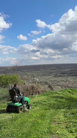 A man is operating a green lawnmower on a grassy hill overlooking a vast valley landscape. The man is wearing a dark shirt and a hat. A wire fence and brush are visible to the left of the lawnmower. The valley below contains sparse trees and what appears to be a road. The sky is bright blue with puffy white clouds. The video is shot from a stationary position.