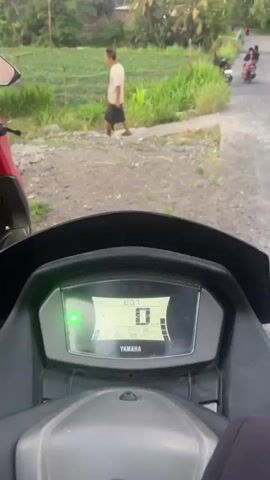 This is a shaky, low-resolution video taken from the perspective of someone riding a motorcycle. The rider, wearing a black hoodie, sunglasses, and a blue helmet, is looking into the camera. In the background, there's a rural landscape with lush greenery, palm trees, and a field. The sky is a pale orange and blue, suggesting either sunrise or sunset. A person is walking in the field, and another motorcycle is visible in the distance on the road. The video appears to be taken in motion, indicated by the blurriness and the movement of the camera.