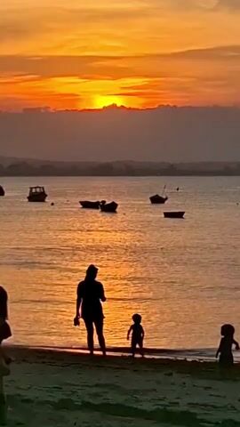 A picturesque sunset illuminates a tranquil scene over a body of water in Armação Dos Búzios, Brazil. The sky is awash with brilliant shades of orange and yellow, with the sun glowing brightly just above a low band of clouds on the horizon. The warm, golden light casts a shimmering reflection across the calm surface of the water.

In the foreground, silhouetted figures stand on the sandy beach. A woman is positioned centrally, with two small children near her to the right; one child appears to be walking towards the water's edge, and the other is closer to the shore. Part of another person is visible on the far left of the frame.

Several small boats are gently floating, anchored across the water, their dark shapes contrasting against the bright reflections. Distant land or islands can be seen faintly across the expanse of water. The overall ambiance is one of peaceful beauty, capturing a serene moment as day transitions to night.