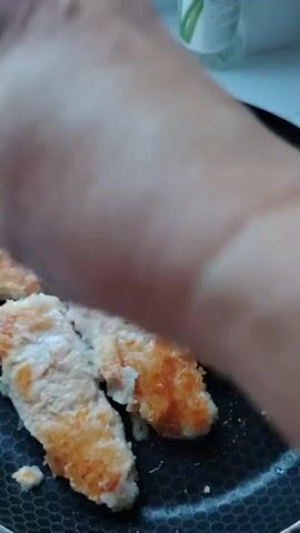A close-up, blurry shot focuses on a hand, likely of an adult, preparing food. The hand is positioned over a circular object, possibly a pan or plate, which contains a light-colored, circular food item that appears to be spread with an orange substance, possibly jam or a sauce. The hand is reaching in and touching the food item, suggesting it's being manipulated or seasoned. The background is out of focus but hints at an indoor setting with hints of white and possibly green. The lighting is soft and diffuse. The video seems to be a short, informal recording of a cooking or food preparation activity.
