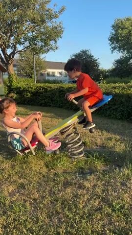 A young girl and boy are playing on a seesaw in a park. The girl is on the left side of the seesaw, wearing a light pink tank top and colorful shorts. She has her hair in a ponytail and is wearing pink sneakers. The boy is on the right side of the seesaw, wearing an orange t-shirt and red shorts. He is wearing black sneakers. The seesaw is yellow and blue and has a metal spring mechanism underneath. The park has green grass and trees in the background. The sky is clear and blue. The children are both holding onto the handles of the seesaw and appear to be enjoying themselves. The video is shot from a slightly low angle, looking up at the children.