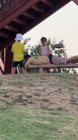 In this video, a man is lying face down on a wooden bench. A young boy in a yellow t-shirt and a white hat is standing to the left of the bench, interacting with the man's head. A young girl in a pink tank top is standing behind the man, also interacting with his head. The scene appears to be outdoors, possibly in a park or recreational area, with trees and dry grass visible in the background. The lighting suggests it is either early morning or late afternoon. The overall mood seems playful and relaxed, with the children engaging in what appears to be a game or a playful interaction with the man. There is no visible text in the video. The location is identified as Geullay Taiman, Israel.