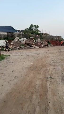 The video shows an outdoor scene in what appears to be a dirt parking lot or undeveloped area. There is a pile of large rocks and dirt against a brick wall in the background. A man walks from left to right in front of the rocks. The ground is uneven and scattered with debris. The sky is overcast.