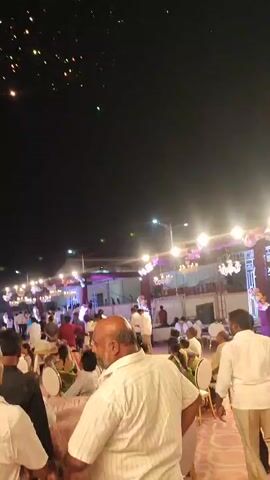 The video captures an outdoor nighttime event in Askihala, India, likely a celebration or gathering. The initial shot shows numerous people seated at tables under artificial lighting, suggesting a festive occasion. The sky above them is dark, with scattered small lights, possibly from earlier fireworks or decorative elements. The scene then shifts to focus on a vibrant fireworks display erupting in the dark night sky. Large, bright bursts of light and sparks fill the frame, accompanied by the sounds of the explosions. The fireworks are the main attraction, illuminating the sky and adding to the celebratory atmosphere of the gathering below.