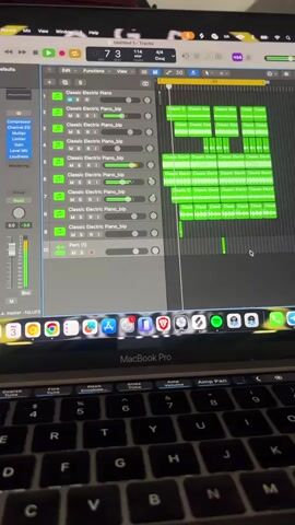 The video displays a close-up of a MacBook Pro laptop screen running Apple's Logic Pro X music production software. The software interface shows a project with multiple audio tracks, predominantly labeled "Classic Electric Piano_blp," along with a single track labeled "Perc (1)." The music is actively playing, indicated by a green playback bar moving across the tracks and fluctuating volume meters on the left side of the screen.

The timecode visible on the screen reads approximately 00:00:09:03, with the project set at a tempo of 114 BPM, a 4/4 time signature, and in the key of C major. The audio features an electronic piano melody accompanied by percussion.

Below the screen, the black keyboard of the MacBook Pro is visible, with the "MacBook Pro" branding centered beneath the display. The laptop's dock at the bottom of the screen displays numerous application icons, including Finder, Safari, Messages, Mail, Logic Pro X, Ableton Live, Adobe Premiere Pro, Adobe Photoshop, Spotify, Google Chrome, and Slack. The scene depicts an indoor setting where music is actively being produced or reviewed.