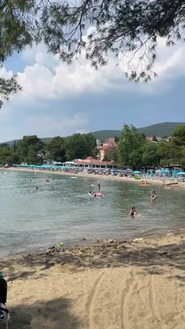 This media file captures a vibrant beach scene in Crikvenica, Croatia. The setting is a sandy beach bordering a calm, clear body of water. The water is populated with numerous individuals enjoying various recreational activities. Some are swimming, while others are relaxing on inflatable rafts. A few people are visible on the shore, and further back, a long row of beach umbrellas suggests a well-established tourist area.

Behind the beach, a line of trees and buildings forms the backdrop. The architecture appears to be Mediterranean in style, with red-tiled roofs visible. Lush green trees dot the landscape, and a gentle slope or hill rises behind the buildings, suggesting the town is situated against a natural incline. The sky above is partly cloudy, indicating fair weather for a summer day, likely mid-afternoon given the bright lighting and activity levels.

The overall mood is one of leisure and enjoyment, characteristic of a holiday destination. The scattered presence of beach toys in the shallow water, like a green bucket, adds to the sense of family fun and relaxation. The framing of the shot with pine tree branches in the foreground adds a natural, airy feel to the composition. No visible text is present in the media file.