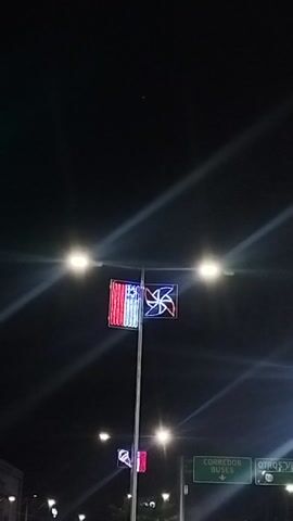 The video captures a nighttime street scene in Maipú, Chile. Two prominent decorative light displays hang from a pole; one is a rectangular design featuring red and white stripes with a blue square and white star, resembling the Chilean flag. Above it, another light display glows in a blue and white pinwheel pattern with red accents. Bright street lights illuminate the foreground, creating a glare on the lens.

In the background, out-of-focus vehicle lights suggest traffic, and green traffic lights are visible. A green road sign indicates "CORREDOR BUSES" (Bus Corridor) and "OTROS. VIA" (Other Routes), providing a location cue.

A man wearing a dark jacket walks into the frame from the right side, pauses briefly to look towards the left, and then continues walking out of view to the left. The ground he walks on appears to be a paved area or sidewalk next to a road. The audio features a person saying "Oh, Chile!", acknowledging the flag display. The overall setting is an urban environment illuminated by artificial lights.