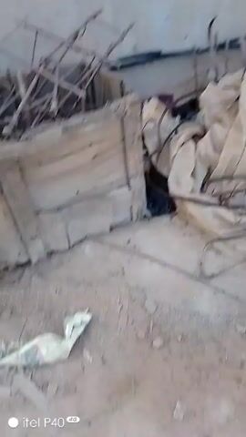 The video shows a cluttered outdoor space littered with construction materials. There are wooden planks, pipes, a red barrel, and what appears to be scrap metal. The ground is dirt, and there's a white wall visible in the background at the start. The camera pans around, offering a wider view of the scattered materials. There are piles of stacked wood and metal structures farther away.