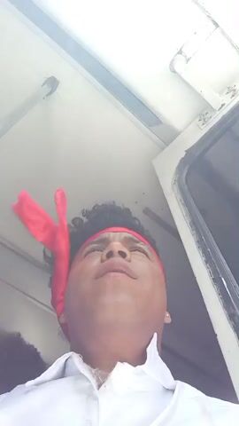 A low-angle, slightly blurry video shows a person with dark, curly hair and a red bandana around their forehead. They are wearing a white collared shirt. The person is looking up and to the left, their mouth slightly open, and then looks directly at the camera with a neutral expression. The background appears to be the interior of a vehicle, possibly a bus, with a white ceiling and a window frame visible. The lighting is bright and appears to be natural. The video captures a close-up of the person's face and upper body.