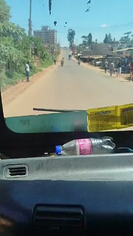 From the interior of a vehicle, a road runs through a town on a sunny day. A water bottle rests on the dashboard, obscuring the view somewhat. The road is mostly empty, with a few pedestrians and motorcycle riders visible in the distance. Buildings and trees line the street, and power lines stretch overhead.