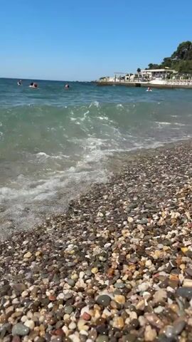The video shows a close-up of a pebbly beach with gentle waves washing ashore. In the background, the clear blue ocean stretches to the horizon under a bright blue sky. Several people can be seen swimming or floating in the water. Along the coastline, there are some buildings, possibly hotels or restaurants, with umbrellas and chairs set up on the beach. A green, tree-covered hill rises behind the buildings. The focus is on the movement of the water and the texture of the pebbles on the beach.