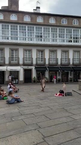 A street performer is dancing in a plaza. The performer is wearing a light-colored dress and a grey jacket. They are standing on a stone plaza in front of a building with white walls and several floors. The second floor has a long balcony with many windows. Some people are sitting on the ground watching the performance. There are a few small businesses on the ground floor of the building. The sky is overcast.