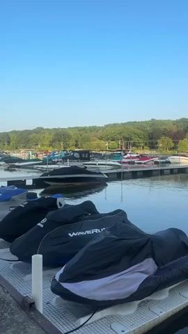 This is a vertical video of a marina with several jet skis parked on a dock in the foreground. The jet skis are covered with dark-colored tarps, some with visible branding such as "WAVERUNNER" and "YAMAHA". In the background, there are several boats docked in the water, also covered or partially covered. The water is calm, reflecting the clear blue sky and the surrounding trees. The environment appears to be a recreational area, likely during the daytime, given the clear sky and bright lighting. There are no people visible in the video.