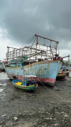 The video shows a large, light blue and pink fishing boat grounded on a muddy shoreline. A smaller green and yellow boat is tied to the larger boat. The sky is overcast with dark clouds. The shoreline is covered in mud and debris. Other boats are visible in the background. There are no people visible in the video. The scene appears to be a harbor or fishing port.