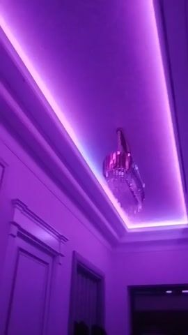 The video presents a low-angle view of an interior space, possibly a room, bathed in vibrant purple light. The walls are white and feature decorative molding. A chandelier, composed of reflective materials, hangs from the ceiling. The ceiling itself is adorned with recessed lighting, creating a bright perimeter of purple illumination. A doorway is partially visible on the left side of the frame, and a window with blinds can be seen below the chandelier. The camera slowly rotates, emphasizing the interplay of light and shadow on the objects and surfaces within the room.