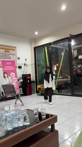 In an indoor waiting area, likely a clinic or salon in Kaliwates, Indonesia, a young girl wearing a black hijab, white long-sleeved top, and black pants walks forward, smiles, and then pauses as an unseen person's hand adjusts her hijab. The area features white tiled floors and bright artificial lighting.

To the left, a pink promotional sign for "CareNow" is visible, advertising dental calculus cleaning ("BERSHKAN KARANG GIGI") and installment payment options ("Cici Biaya Perawatanmu") with details like "0%" and "50rb". Below the sign are some waiting chairs. In the background, behind the girl, a yellow generator, a red and black water dispenser, and a black trash can are seen. Beyond glass doors, a "TUTUP" (CLOSED) sign is displayed, with a car and motorbikes visible outside. In the foreground, a brown table holds numerous empty and some full glasses of water, along with a smartphone. The girl appears happy and cheerful.