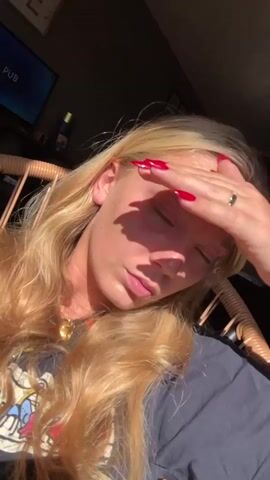 A woman with long blond hair and fair skin is sitting in a wicker chair. She is wearing a dark t-shirt and a gold necklace. Her nails are painted red. The sun is shining on her face, and she is holding her hand up to block the light. She sticks her tongue out.