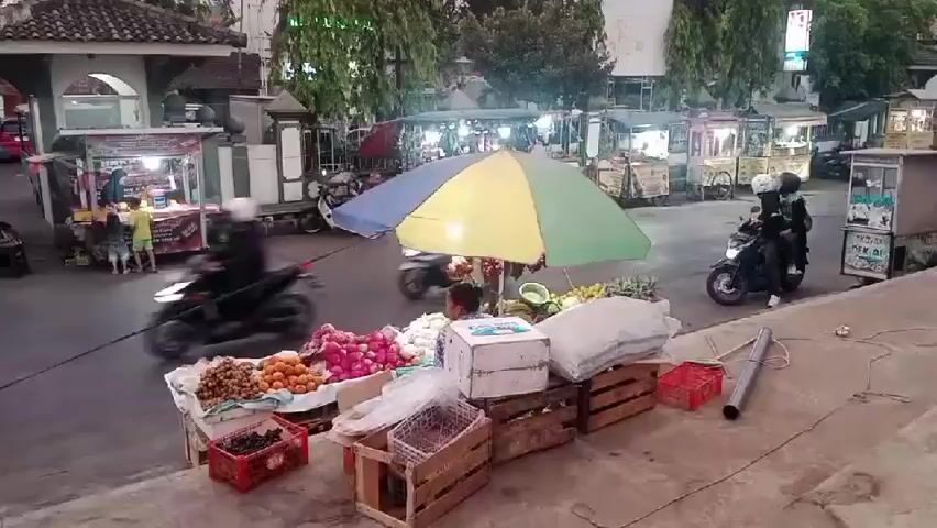 A street market scene is depicted at dusk. In the foreground, a fruit stand is set up on the sidewalk, displaying an assortment of fruits such as grapes, oranges, and what appear to be apples or tomatoes. The stall is covered by a colorful umbrella, and several wooden crates are used as display platforms. A person is seated behind the stall, partially obscured by the produce. To the right, a motorcycle with two riders passes by, and further back, another motorcycle is parked. In the background, several food stalls are visible, with lights illuminating them. People are seen interacting at these stalls. The street itself is paved, and there are trees lining the area. The lighting suggests it is either early morning or late evening.