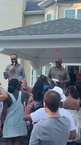 A man on a stage holds a microphone and speaks to an audience. He wears sunglasses, a patterned button-down shirt, and dark pants. Another man stands to his right, holding a white cloth and gesturing with his hands. He wears a collared shirt and shorts. The audience members, many holding up phones to record, are visible in the foreground. The scene appears to be an outdoor event, possibly a party or gathering, with a house in the background and what looks like a covered patio area above the stage. The lighting suggests it is late afternoon or early evening.