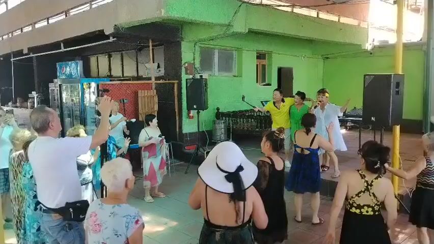 A group of adults are gathered in what appears to be an outdoor covered area, possibly a restaurant or bar, with green walls. Several people are dancing, while others stand and watch. In the center of the scene, a group of five individuals are singing and performing, with one person holding a microphone and another a selfie stick. Two large speakers are positioned on either side of the performing group. In the background, there are refrigerators, shelves, and a large wooden structure. The lighting suggests it is daytime.