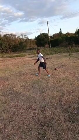 A person wearing a white t-shirt, dark shorts, and light blue sandals is seen standing in a dry, grassy field. The person starts to walk away from the camera, then begins to run towards the left side of the frame. The background consists of trees, bushes, a power line, and a sky with scattered clouds.