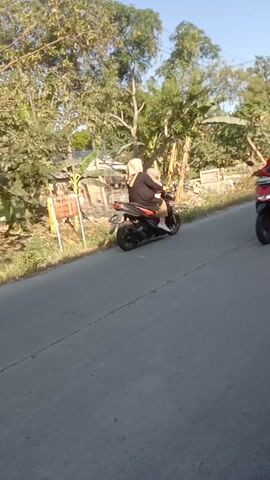 Two people are riding on a red motorcycle on a paved road. The person in front is wearing a helmet and a jacket. The person in the back is wearing a headscarf and a light-colored garment. The motorcycle is moving forward. The environment appears to be outdoors with trees and vegetation visible on the side of the road. Another black motorcycle is parked in the foreground, partially obstructing the view. The sun appears to be shining.