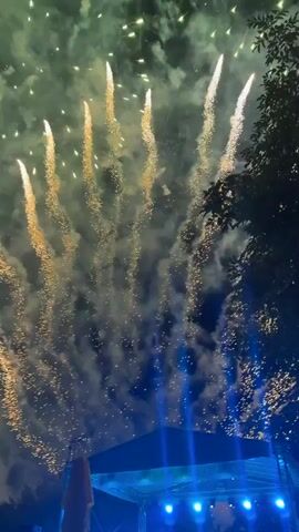 The video shows a fireworks display at night. Multiple fireworks are exploding in the sky, creating bright bursts of light and smoke in various colors, including green, yellow, white, and red. Some fireworks leave trails of sparks as they ascend and descend. In the foreground, a dark silhouette of trees is visible on the right side of the frame, and a dark structure with blue lights is present at the bottom. The scene suggests an outdoor event or celebration.