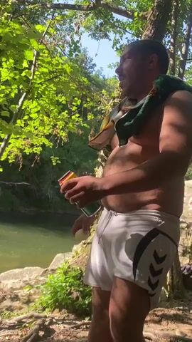 The video shows a man standing near a body of water, possibly a river, in a wooded area. He is shirtless, wearing white athletic shorts with a black design on the side. A colorful towel is draped over his shoulders. He holds a can in his right hand and a small white object, possibly a ball or egg, in his left. The man is looking to his right and appears to be speaking. The environment is lush with green foliage and sunlight filtering through the trees. The water is visible in the background, along with rocks and dry leaves on the ground.