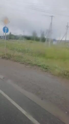 The video shows a side view of a road with some water and mud on it, suggesting it might be raining or has recently rained. There are grass and trees on the side of the road, along with utility poles and wires. Several road signs are visible. One sign is a white diamond with a yellow center, indicating a priority road. Below it is a blue circular sign with a white arrow pointing upwards, signifying a mandatory direction. Further down the road, a blue rectangular sign displays numbers and an arrow pointing to the right. Another blue sign with white arrows indicating traffic direction is also present. The overall environment appears to be rural or suburban, with an overcast sky.
