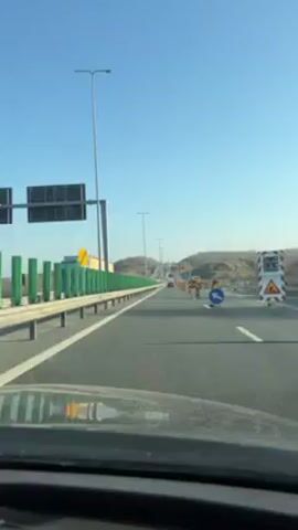 The video shows a car driving on a highway. The sky is blue. The road has multiple lanes and is lined with metal guardrails. There are orange and white striped safety markers and construction vehicles along the right side of the road, indicating construction or road work. The camera view is from inside the car, looking out through the windshield.