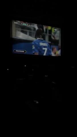 The video shows a large screen displaying a soccer match. On the screen, a soccer player wearing a blue jersey with the number 7 and the name "DEMBELE" on the back is shown. The player is looking to the right. Further down the screen, there are graphics indicating the game's score and time. In the foreground, two men are watching the screen. The man on the left is bald and looking intently at the screen. The man on the right is wearing glasses and also looking at the screen. The environment is dark, suggesting the video is being watched in a theater or a room with the lights dimmed. There are faint lights visible in the darkness below the screen. The screen itself has a border and the displayed image is bright.