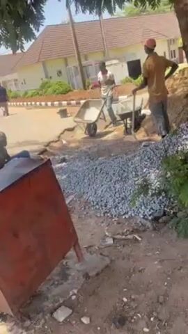 In a medium shot, three construction workers are working on a street in a residential area. Two of the workers are standing by a pile of gray gravel, with one leaning on a shovel and the other holding a wheelbarrow loaded with the gravel. The third worker is sitting on the ground, next to a red metal box. The street in front of them is cracked and broken, and in the background, there are light-colored houses with brown tile roofs. Trees provide shade over the area. The workers are wearing casual clothes and hats, and their faces are visible. Another figure is walking in the background on the left side of the frame. The camera is steady.