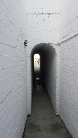 A person walks through a narrow, white-walled arched tunnel. At the end of the tunnel, the view opens up to a sunny beach scene with the sea, rocks, beach chairs, and an umbrella visible.