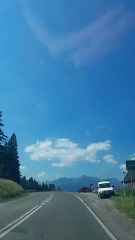 The video shows a view from inside a vehicle driving on a paved road. The road curves to the left and then to the right. On the right side of the road, there is a red car parked near a wooden signpost. In the distance, mountains are visible under a clear blue sky with scattered white clouds. On the left side of the road, there are trees and green grass. The weather appears to be sunny and clear.