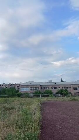 The video displays an outdoor scene, panning from left to right across a dirt field or track. The foreground features patchy green grass and low vegetation along the left edge of the field. In the midground, several buildings are visible. Initially, a long, low-rise building with numerous windows, possibly a school or administrative building, stands behind a line of trees. As the camera pans, more multi-story residential buildings, typical of post-Soviet architecture, come into view in the background, with multiple windows and balconies. A white football goalpost is briefly seen on the right side of the dirt field. Overhead wires or power lines stretch across the frame against a sky filled with a mix of white clouds and blue patches. The lighting suggests it is daytime, possibly late afternoon, under partly cloudy weather. No people or specific activities are visible in the scene. The architecture is consistent with urban environments in places like Vagharshapat, Armenia. No discernible text is visible on any objects or buildings.