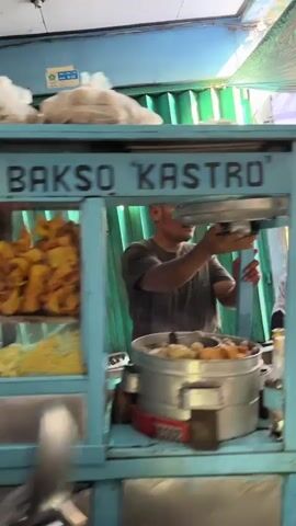 A man is shown working at a food stall. The stall is blue and has the word "KASTRO" written on it. The man is wearing a dark t-shirt and dark pants. He is in the process of preparing food, as evidenced by the steaming pot in front of him. The background shows a green corrugated metal wall and some pipes. The lighting suggests it is daytime.