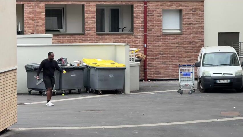 In an outdoor urban area, a man in a black long-sleeved shirt, black shorts, and white sneakers is hitting a tennis ball with a tennis racket. He is standing in a paved area next to a row of gray trash bins. A white delivery van is parked on the right side of the frame, and a shopping cart is standing in front of it. The background includes a brick building with windows.