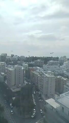 The video shows a high-angle, daytime view of an urban landscape. The sky is overcast and gray, with a flat, even cloud cover. Below, a dense collection of buildings, mostly gray and white in color, stretches toward the horizon. A main road bisects the foreground, with vehicles moving along it and parked on the sides. In the distance, the sea is visible, with several ships or boats dotting the horizon. On the left side of the frame, two tall, modern skyscrapers rise above the other buildings. The overall impression is of a densely populated city under a cloudy sky.