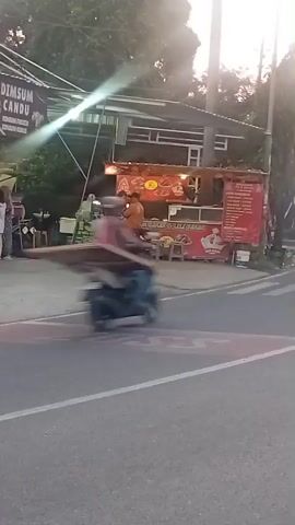The video shows a street scene with several food stalls. On the left, there is a stall with a sign that says "DIMSUM CANDU". Two people are standing in front of it. To the right, there is a larger food stall with a red awning and various dishes displayed. Several people are visible near this stall. Two motorcycles are present, one is parked and another is moving across the street. The overall environment appears to be a bustling street with food vendors. The lighting suggests it might be late afternoon or early evening.