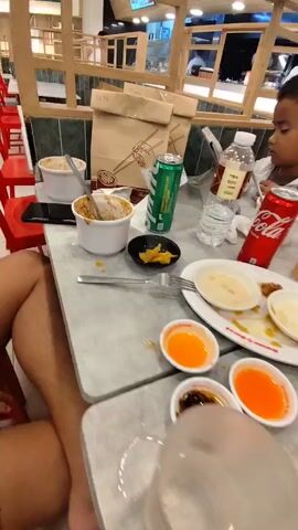 The video shows a table in a restaurant with the remnants of a meal. There are several plates with food scraps, small bowls of dipping sauce, empty cups, and used utensils scattered across the table. A young child is seated at the table, looking away from the camera. Various food and drink containers, including cans of soda and bottles of water, are also visible. The environment appears to be a casual dining establishment with other tables and seating in the background.