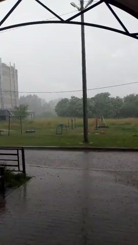The video shows a courtyard during a heavy rainstorm. The ground and pavement are wet, with visible raindrops hitting the surface. In the background, a playground with swings and climbing structures is visible, along with trees and a building under construction. The sky is overcast and gray. The camera appears to be positioned under a covered area, possibly a porch or awning, looking out at the scene. No people are visible in the frame. The overall atmosphere is somber and wet due to the downpour.