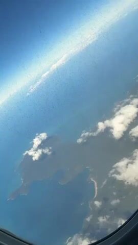 An aerial view from an airplane window shows a vast expanse of blue ocean, a landmass, and scattered white clouds. The landmass, possibly an island or coastal region, is predominantly green with some darker areas, and a visible sandy beach curves along its edge. The ocean displays varying shades of blue, with some lighter patches. Several white, puffy clouds drift over both the land and the sea. The sky above is bright blue, with a horizontal streak of brighter light in the upper portion. The dark frame of the airplane window is visible at the bottom and right edges of the shot, and the window glass has numerous small specks or dust. The entire scene is captured at an oblique angle.