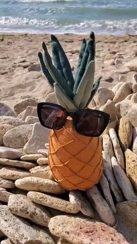 A pineapple with sunglasses sits on a beach. The pineapple is orange and textured to resemble a real pineapple, with green fabric leaves sticking out the top. It is resting on a wall made of small, light-colored rocks. Behind the pineapple, there is a sandy beach with more rocks scattered on it, and the ocean with small waves in the background. The sun is shining, creating reflections in the pineapple's sunglasses. The scene is static, with no discernible movement.