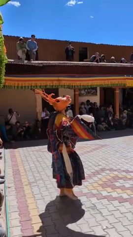 The video opens on a brightly lit courtyard paved with checkered tiles. In the center, a person clad in a vibrant, ornate costume and an animal mask with antlers performs a traditional dance. The mask is orange with pointed, antler-like protrusions. The costume is a riot of colors and patterns, featuring deep blues, reds, yellows, and intricate designs. The dancer holds a white fan in one hand. Behind the dancer, a crowd of spectators is gathered, some seated, others standing, observing the performance. Above the courtyard, a building with a reddish-brown roof is visible, with more people watching from its edge. The sky is a clear, bright blue, indicating a sunny day. The scene suggests a cultural festival or performance taking place in a historic or monastic setting.