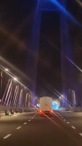 The video shows a white truck driving on a road at night. The truck has its taillights on. The road is lit by streetlights and the headlights of other vehicles. In the background, there is a large bridge with blue lights illuminating its structure. The bridge has tall towers and cables. The camera is moving, suggesting it is also on the road.
