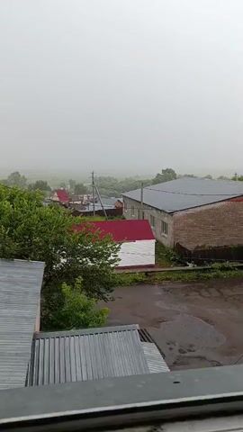 The video shows a view of a rural village on a cloudy day. Rain is visible falling on the roofs of houses and the wet ground. The scene includes several buildings with red and gray roofs, green trees, and power lines. The overall atmosphere is gray and hazy, indicating overcast weather conditions.