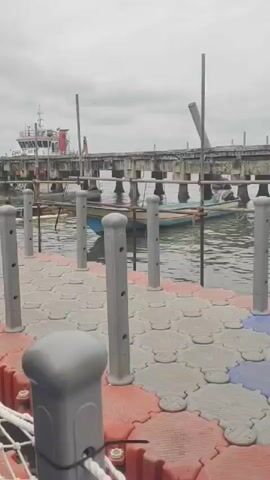 The video shows a person with a black backpack walking on a floating dock. In the background, a large ship and a crane are visible. Other people are also seen walking on the dock with luggage, some of them appearing to be heading towards a larger structure at the end of the dock. The sky is overcast, and the water appears calm. The dock itself is made of interlocking plastic modules, with white railings along the sides.